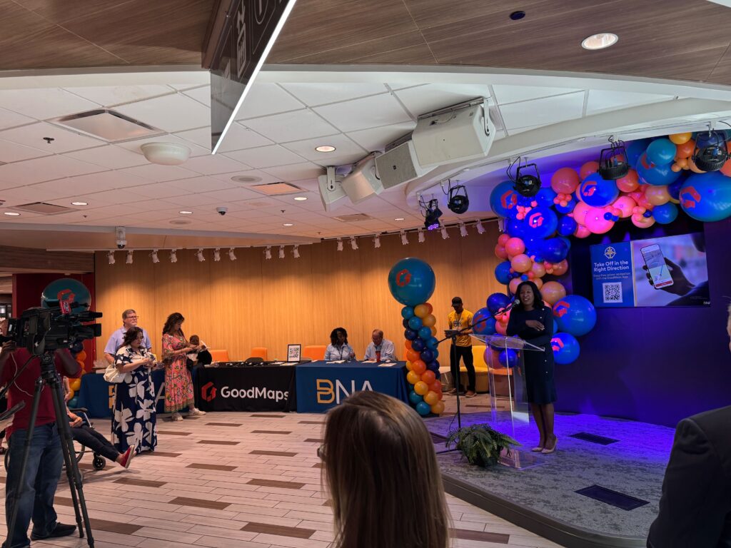 A woman stands at a podium on a stage decorated with colorful balloons, addressing an audience at an indoor event. On the right, a screen displays a smartphone and the text “Take Off in the Right Direction” alongside a QR code. Several people are gathered, some standing and some sitting at tables with “GoodMaps” and “BNA” banners. A cameraman records the scene, while others watch and listen in the brightly lit room.