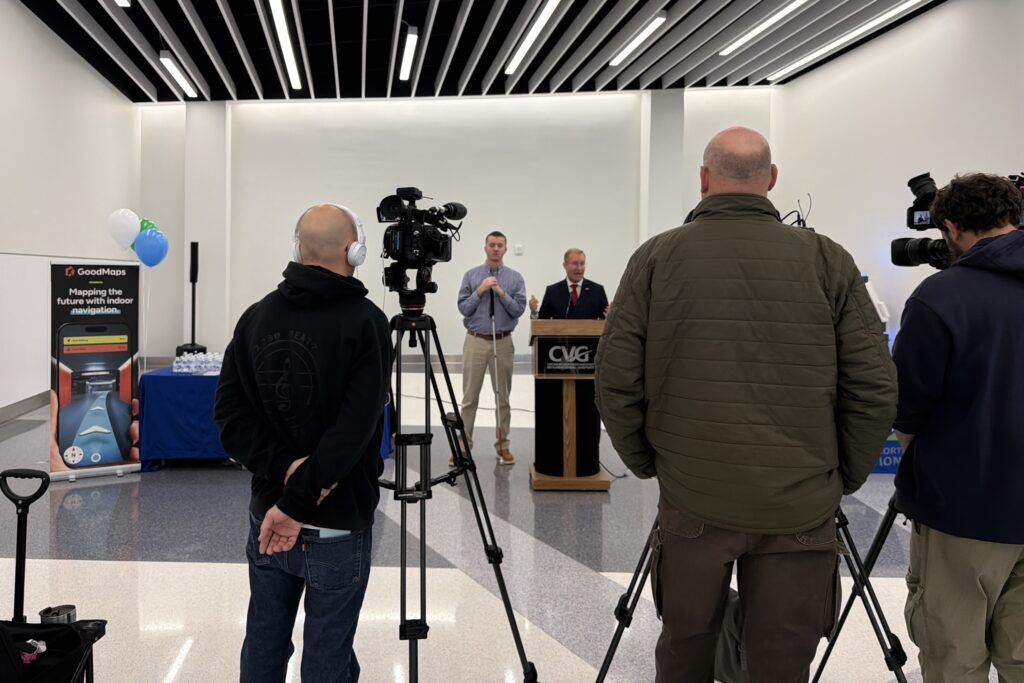 Group of videographers with cameras on tripods film two speakers (one of which is Kevin Kline) standing at a podium during an event in an airport terminal.