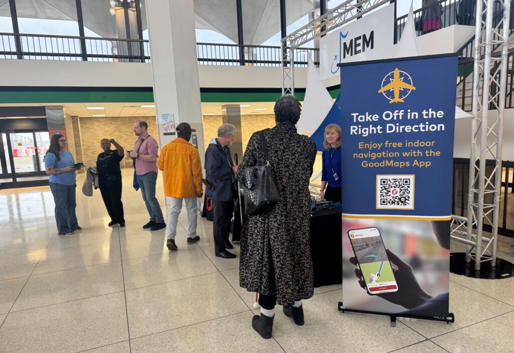 Several travelers gather in an airport concourse near a tall blue GoodMaps promotional banner that reads “Take Off in the Right Direction – Enjoy free indoor navigation with the GoodMaps App,” with a MEM airport sign visible above.