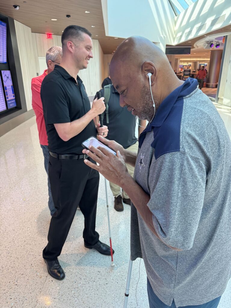 Kevin and several users stand together at BNA. In the foreground, one person holds their smartphone with the GoodMaps app open in one hand and a white cane in the other.