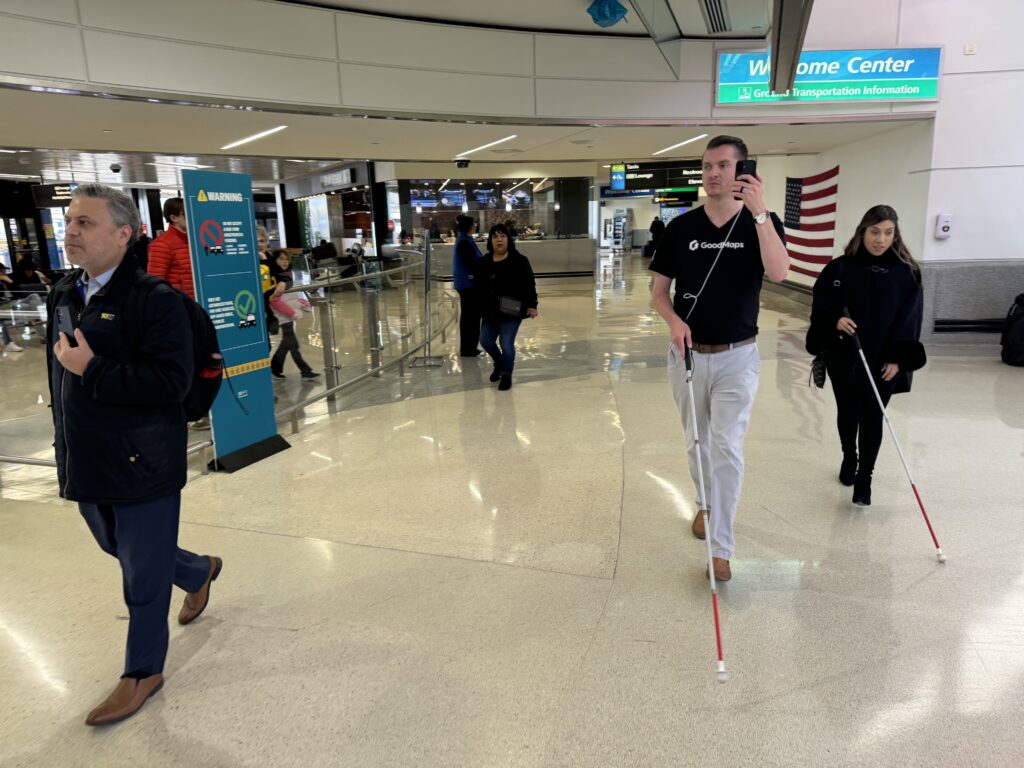 Kevin and Alba walk through the airport holding their smartphones listening to audio directions while using their canes for guidance.