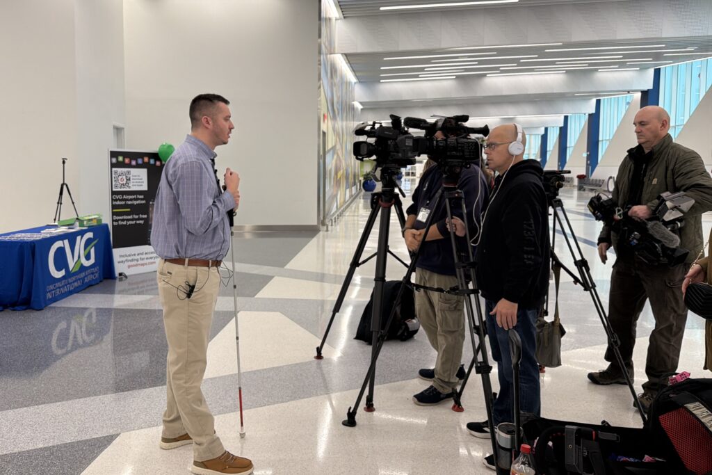 Kevin Kline (GoodMaps Community Engagement Rep) holding his white cane speaks to a group of TV news videographers filming him with cameras on tripods inside an airport concourse.