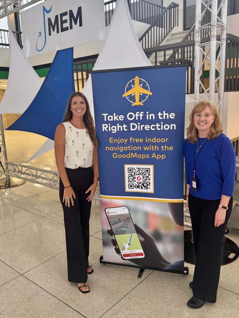 Two women stand beside a blue airport banner that reads “Take Off in the Right Direction – Enjoy free indoor navigation with the GoodMaps App” beneath a MEM airport sign and display structure.