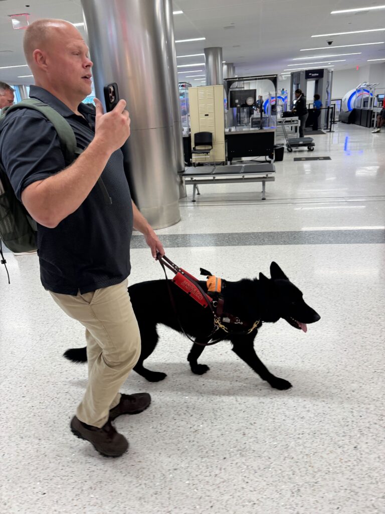 A man holds his smartphone with the GoodMaps app open in one hand and the lead for his guide dog in the other.