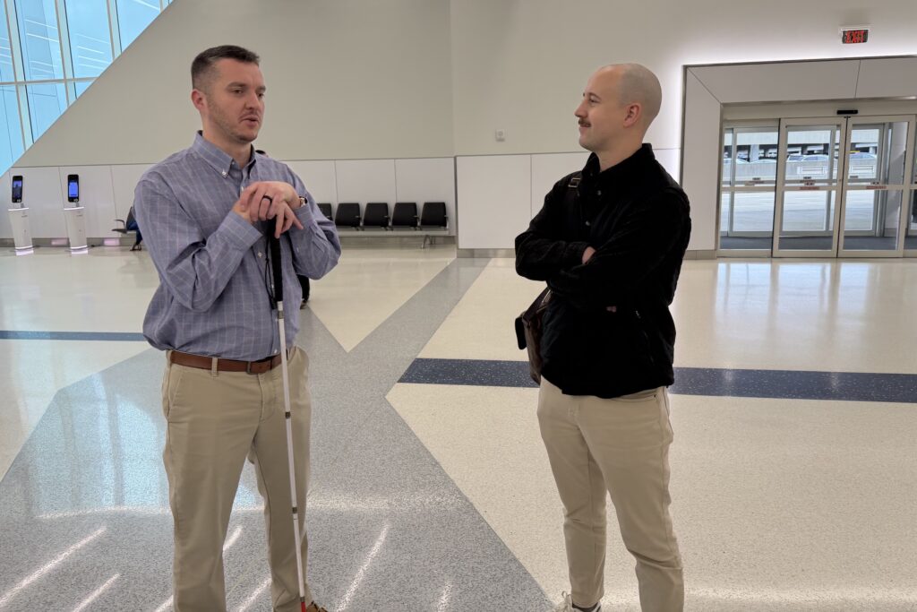 Kevin Kline (GoodMaps Community Engagement Rep) with his white cane talks with another man standing nearby in a mostly empty airport waiting area.