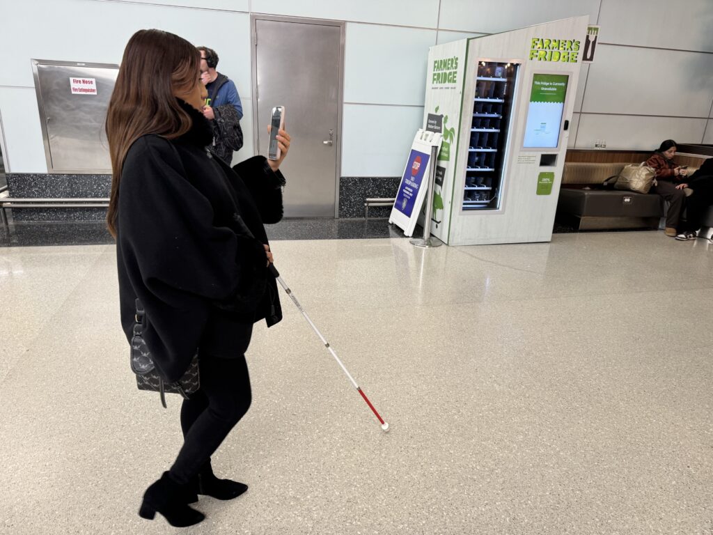 Alba walks through the airport holding her smartphone listening to audio directions while using her cane for guidance.