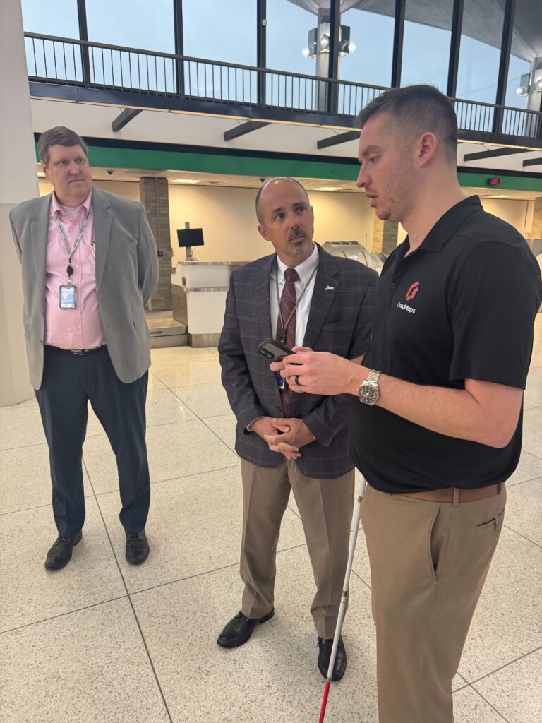 Two men in business attire stand in an airport terminal while a third man in a black GoodMaps polo shirt demonstrates something on a smartphone and holds a white cane.