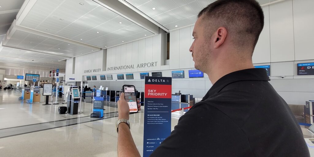 Kevin Kline holds up his smartphone displaying the GoodMaps navigation app inside Lehigh Valley International Airport, standing near a Delta Sky Priority sign and the check-in counters.
