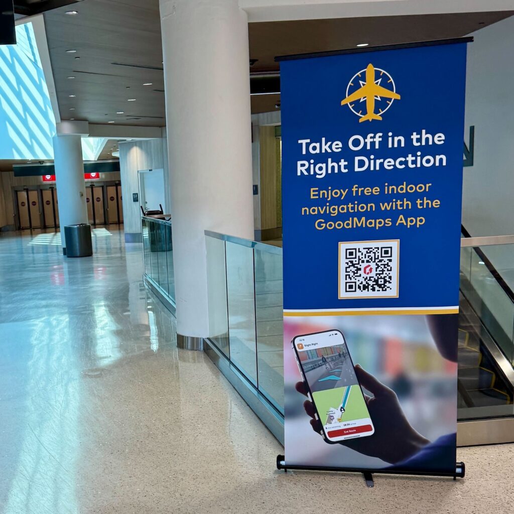 A blue and yellow airport sign advertising the GoodMaps App for free indoor navigation, featuring a QR code and a photo of a hand holding a smartphone displaying a map, located in an airport concourse.