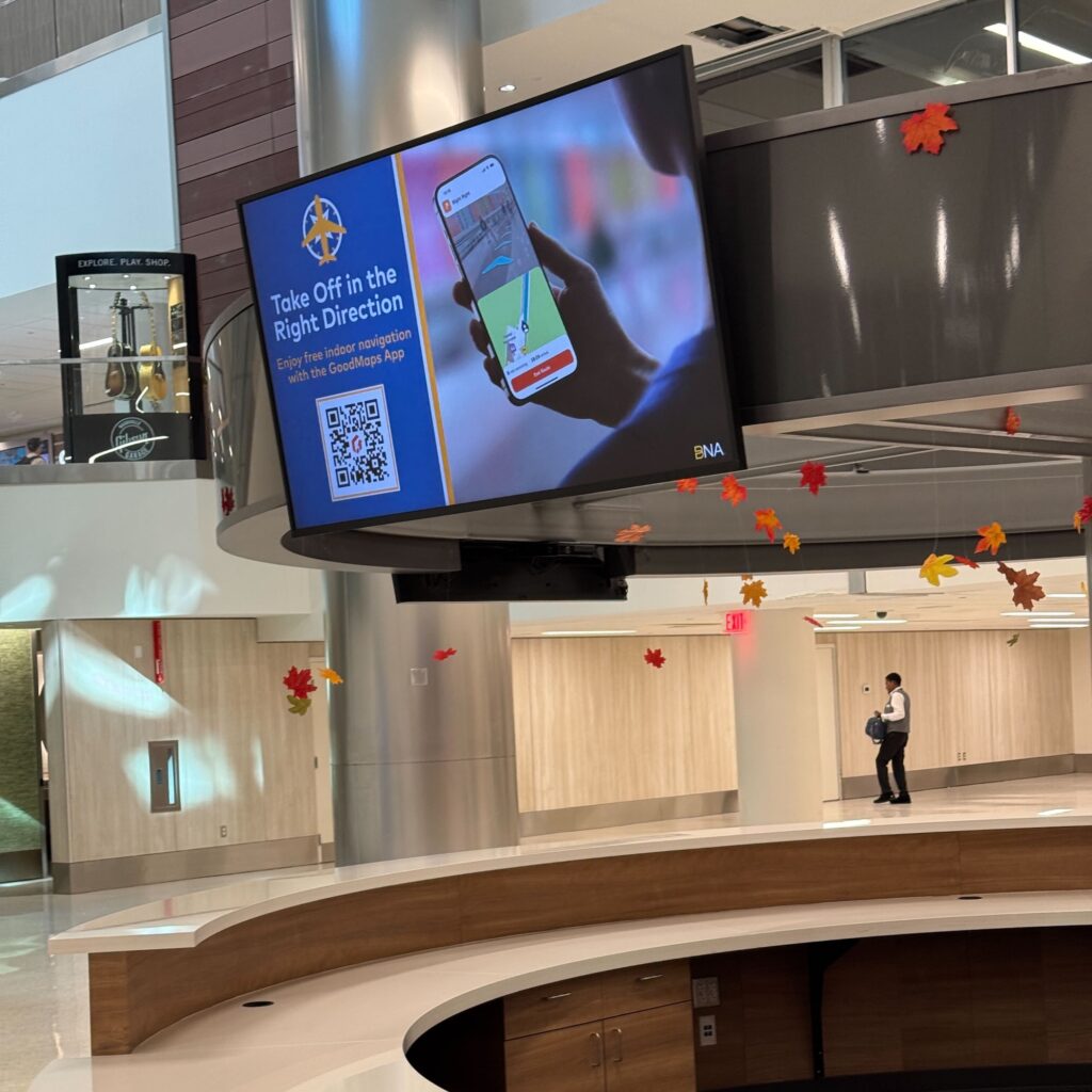 A large electronic screen in an airport displaying an advertisement for the GoodMaps App, showing a hand holding a smartphone with a map; autumn leaf decorations hang from the ceiling nearby and a person walks in the background.