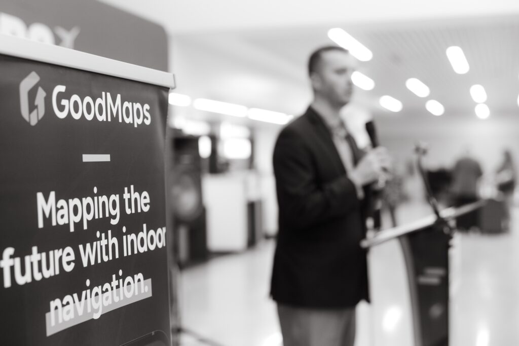 Black‑and‑white photo of a GoodMaps banner reading ‘Mapping the future with indoor navigation’ with a blurred speaker at a podium in the background.