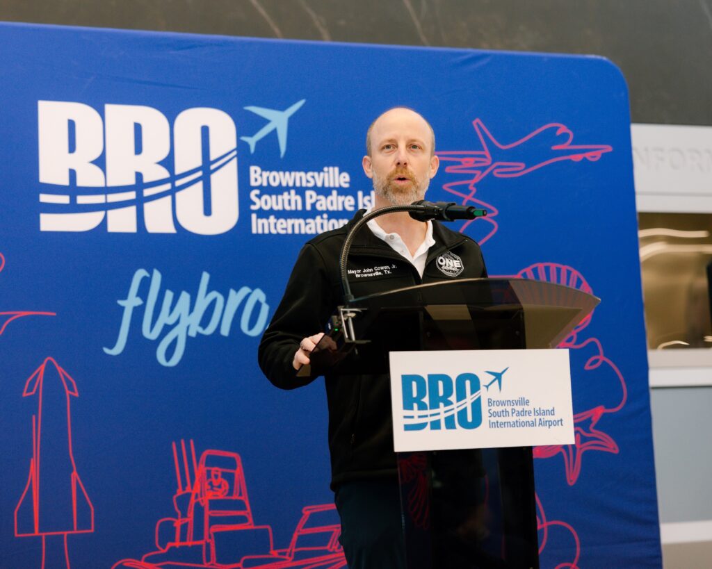 Man speaking at a podium with a microphone in front of a BRO airport backdrop, giving a formal address inside the terminal.