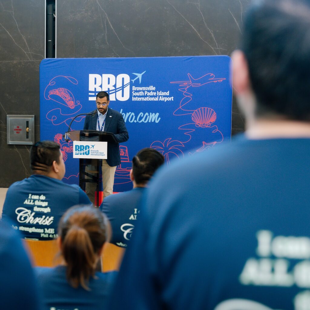 Man in a blazer speaking at a podium in front of a BRO airport backdrop, with an audience in matching blue T‑shirts seated in the foreground.