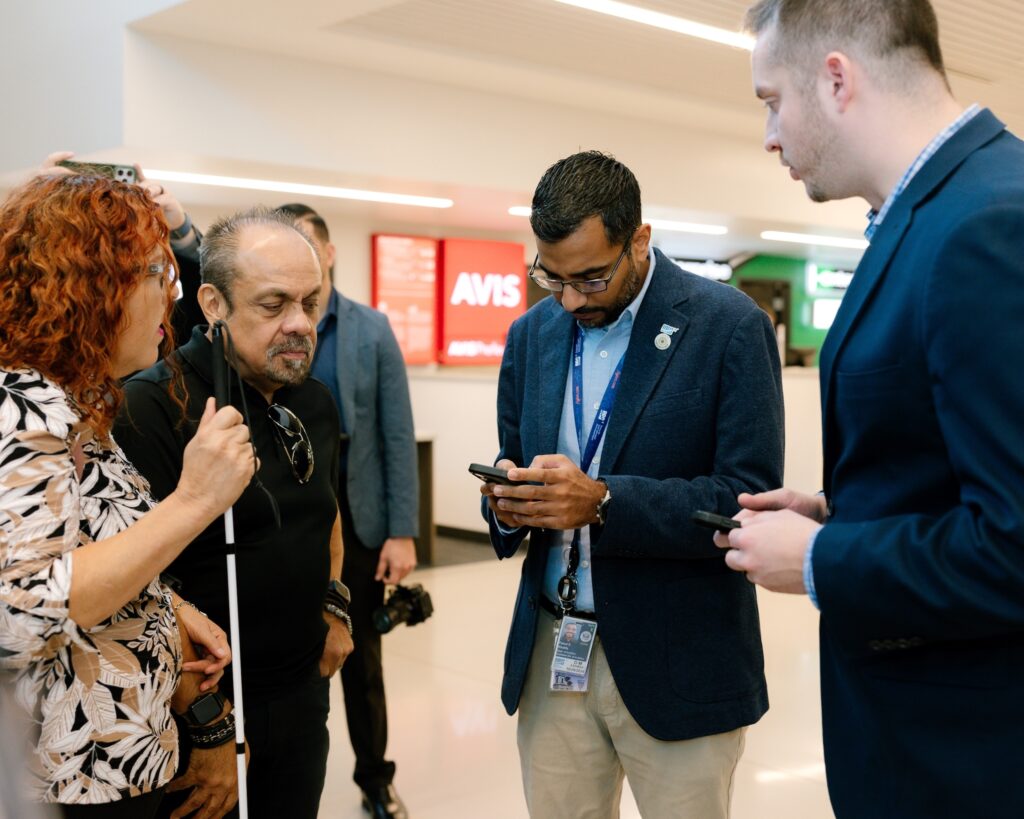 Airport staff member and two guests with white canes standing near rental car counters, looking at smartphones during a demonstration.