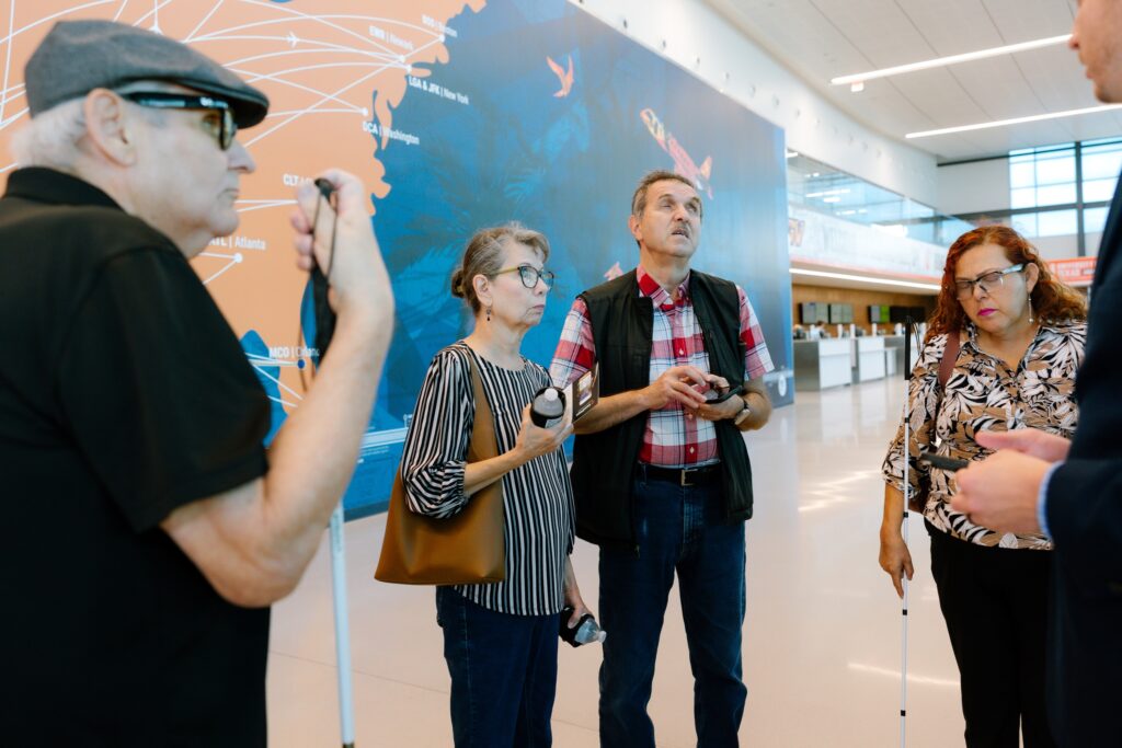 Group of adults with white canes standing in an airport terminal, talking together in front of a large colorful wall map.