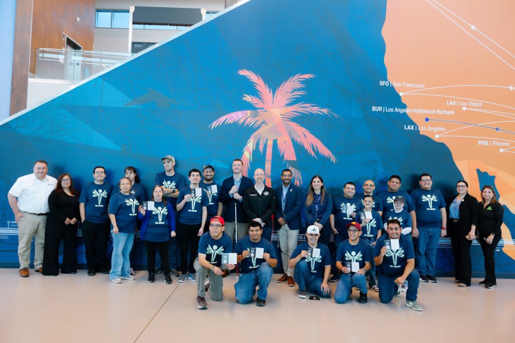 Large group of people posing for a photo in an airport terminal, many wearing matching navy shirts, in front of a blue wall mural with a palm tree and flight routes.