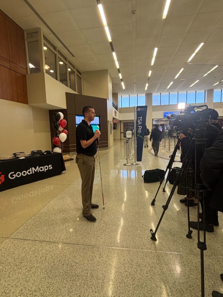 Kevin holds his white cane standing in an airport terminal being interviewed by a camera crew. Behind him, there is a table with a GoodMaps banner and red, black, and white balloons. TSA Pre signage is visible in the background.