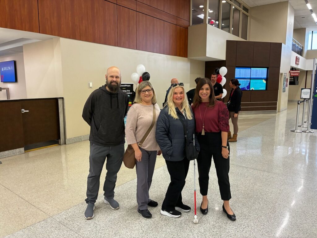 Four people stand together posing for a photo in the LEX terminal. One woman, holding a white cane, is in front, and there are balloons and a GoodMaps banner behind them. Other people are visible in the background near the airport information screens.