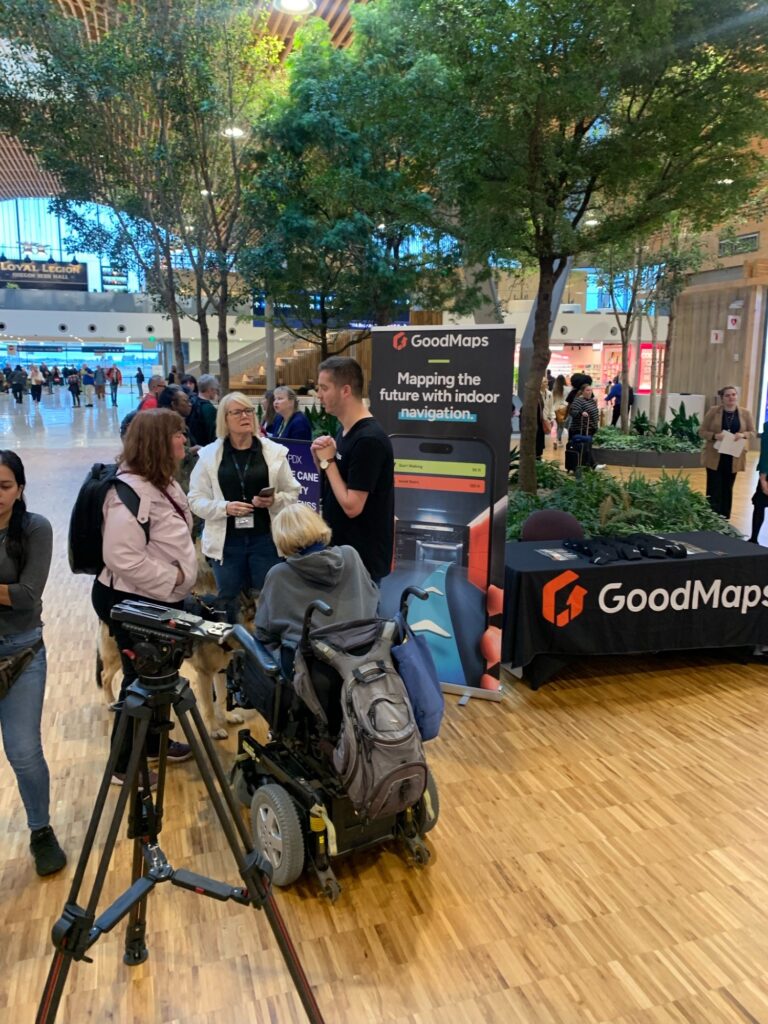 Indoor airport concourse with tall trees and wood flooring where a GoodMaps information booth is set up; a person using a power wheelchair with a service dog and several other people stand talking to a GoodMaps staff member beside a tripod and branded table.