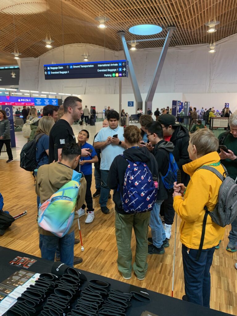 Busy airport arrivals hall with wood flooring where a GoodMaps table is in the foreground and a group of mostly blind or visually impaired travelers with white canes gather around a staff member holding a phone, beneath large overhead directional signs.
