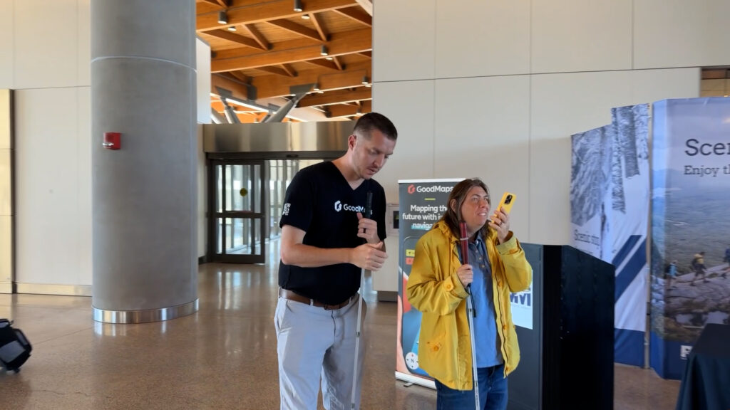 Two people in an airport, one wearing a GoodMaps shirt and holding a white cane, the other in a yellow jacket using a smartphone and white cane, with a GoodMaps banner in the background.