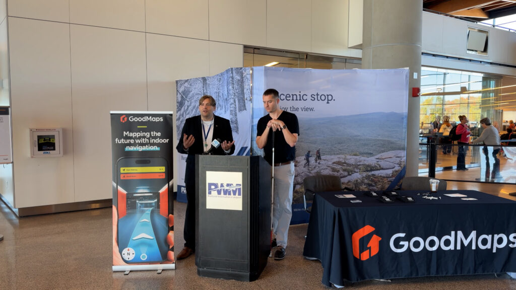 Press event setup at an airport, featuring two speakers at a podium labeled ‘PWM,’ a GoodMaps banner and table, and a scenic promotional display, with people visible in the background.