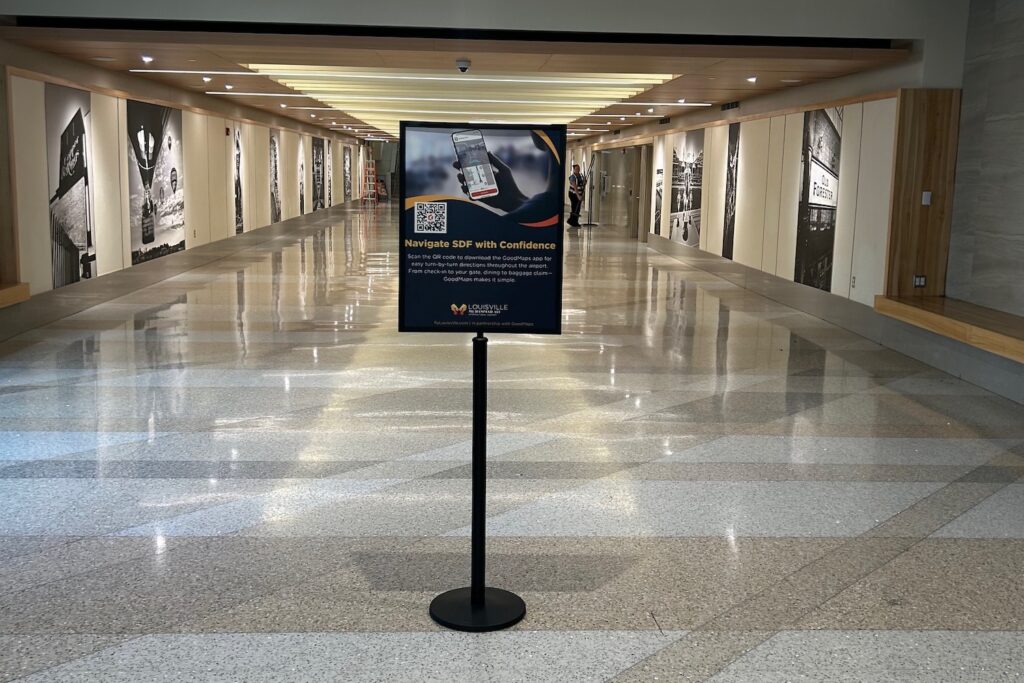 A sign in the center of a polished, empty airport hallway, advertising the GoodMaps app for navigating SDF Airport, with a QR code and a photo of a smartphone on the sign.