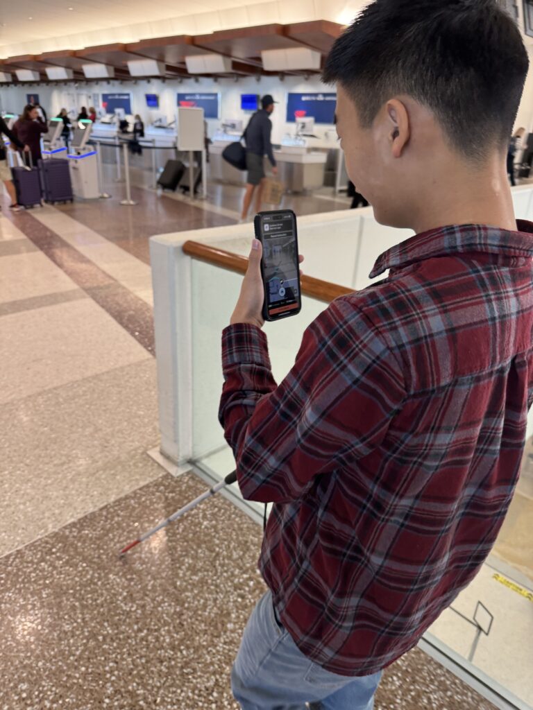 A user holds their smartphone running GoodMaps in one hand and their cane in the other as they navigate SDF.