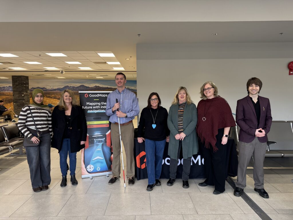Group of eight adults standing in a row inside an airport terminal, posing in front of a GoodMaps accessible navigation display and table, with seating and large landscape photos visible in the background.