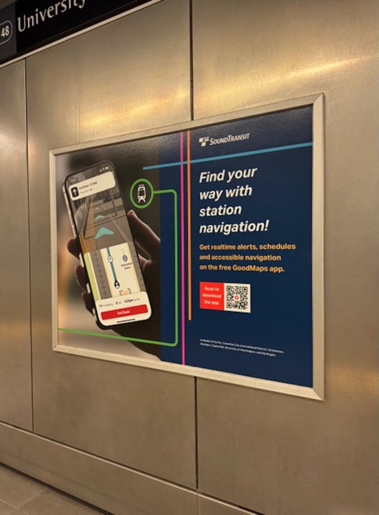 Advertisement poster for SoundTransit inside a subway station, showing a hand holding a smartphone with a navigation app. The poster reads “Find your way with station navigation!” and promotes the free GoodMaps app for real-time alerts, schedules, and accessible navigation, with a QR code for downloading the app. The station name ‘University of Washington’ is partially visible at the top left.