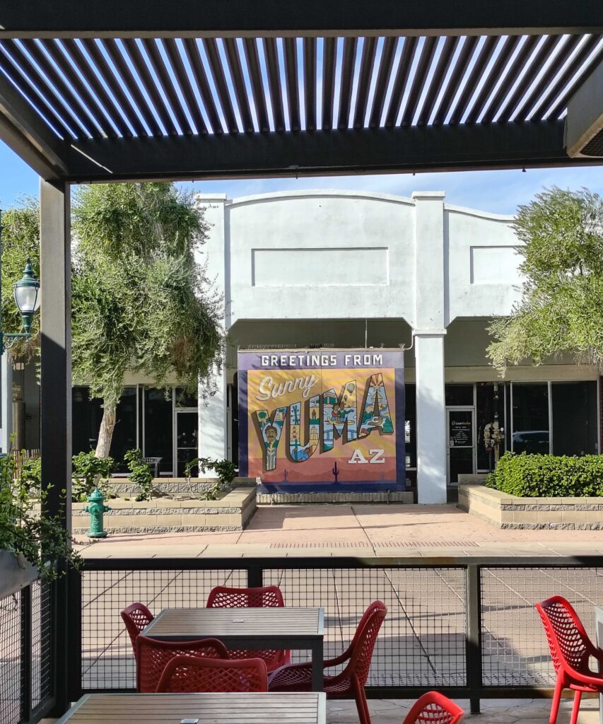 Outdoor mural reading ‘Greetings from Sunny Yuma AZ’ viewed from a patio with red chairs and tables.