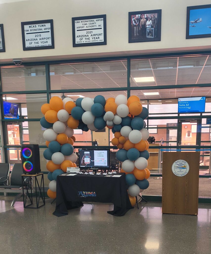 Colorful balloon arch framing an information table and podium inside Yuma International Airport terminal.