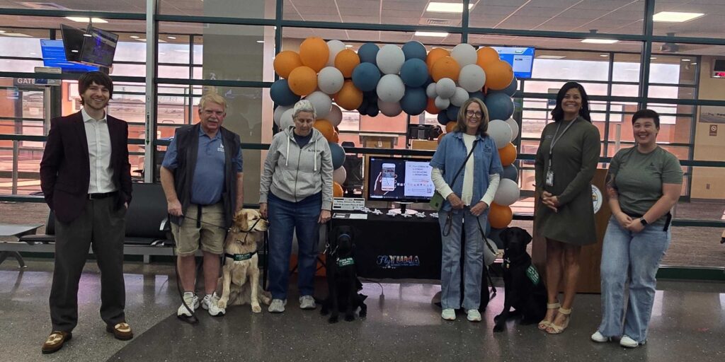Group of people with three service dogs posing in front of a balloon arch and information table inside Yuma airport terminal.