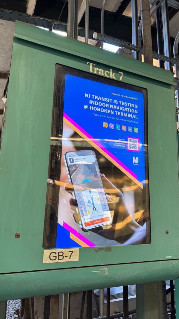 Digital display at Hoboken Terminal Track 7 showing NJ TRANSIT's indoor navigation test announcement with a phone displaying a wayfinding app.