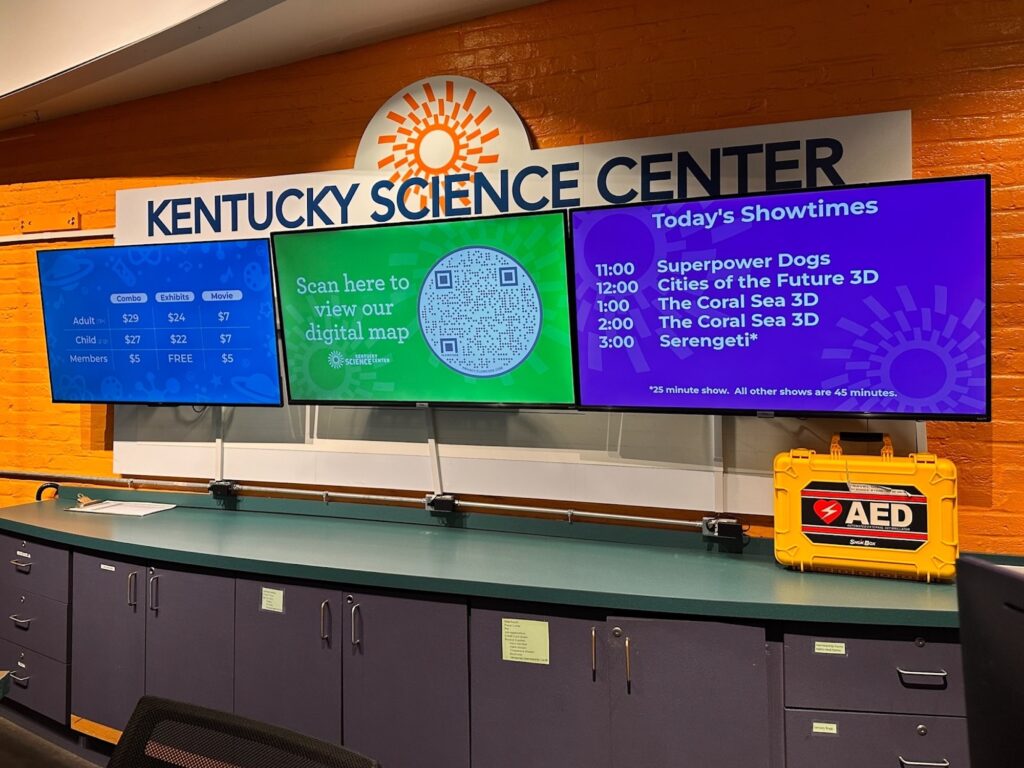 Admission desk at Kentucky Science Center with three digital screens showing ticket prices, a QR code to view the digital map, and a schedule of 4-story theater showtimes.