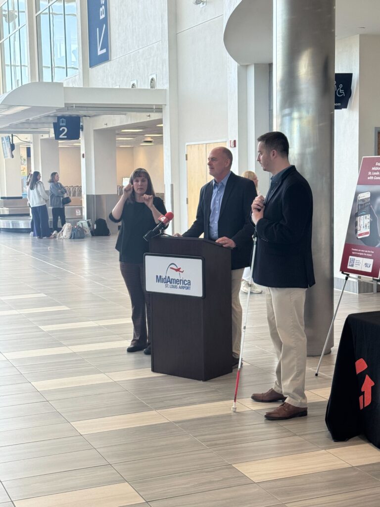 Speaker at a MidAmerica St. Louis Airport podium with a sign language interpreter and Kevin holding his white cane beside the GoodMaps display sign in a bright terminal area.
