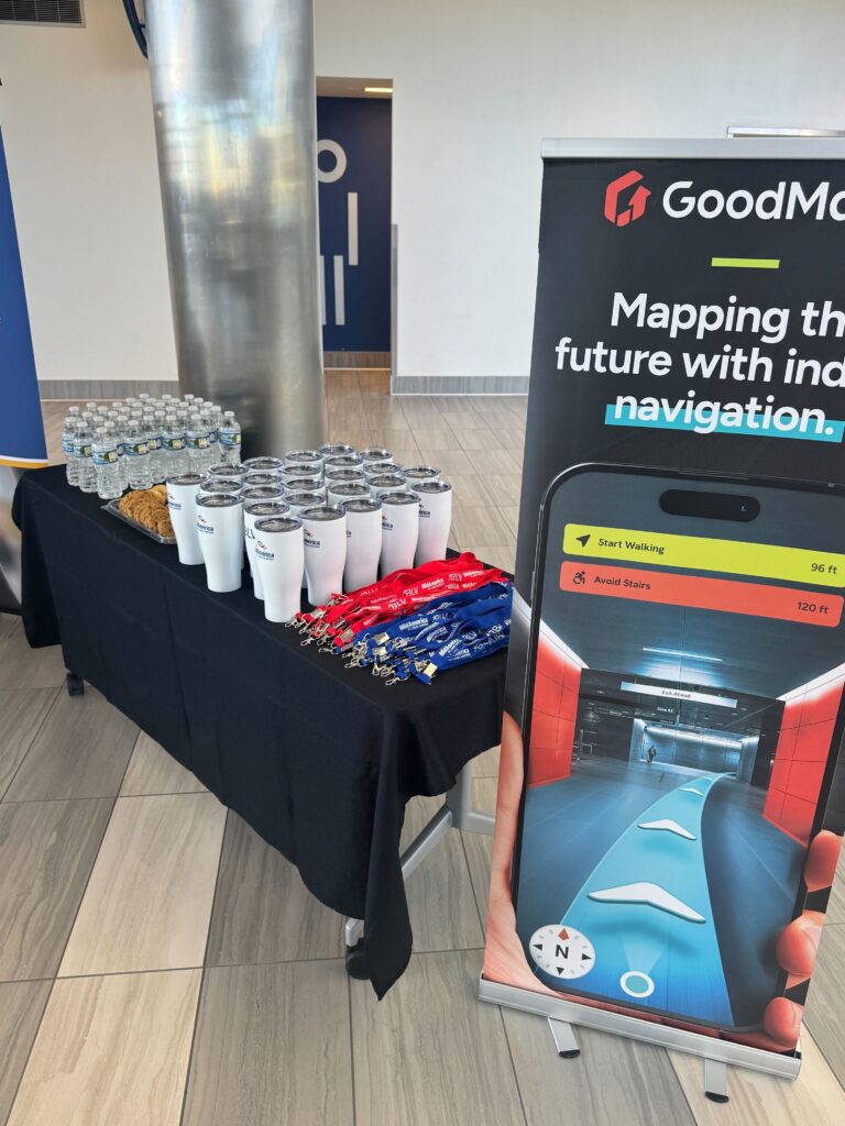 Refreshment table at an event with GoodMaps signage, showing a banner with the navigation app beside a black table with water bottles, branded tumblers, cookies, and lanyards.