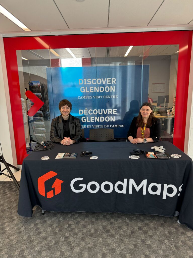 Two people sitting behind a GoodMaps information table at the Discover Glendon Campus Visit Centre, with brochures and promotional items laid out in front of them.