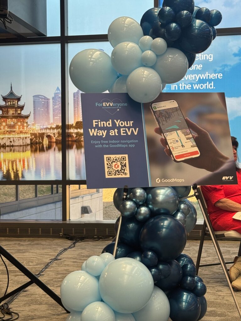 A GoodMaps ‘Find Your Way at EVV’ sign with QR code is mounted on a stand decorated with clusters of light and dark blue balloons inside the airport.