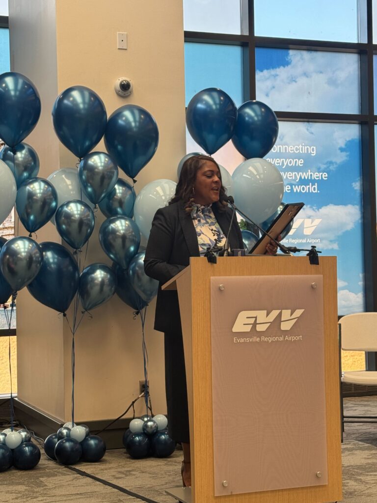 A speaker stands at a podium branded with the Evansville Regional Airport logo, addressing an audience in front of a backdrop of blue balloons.