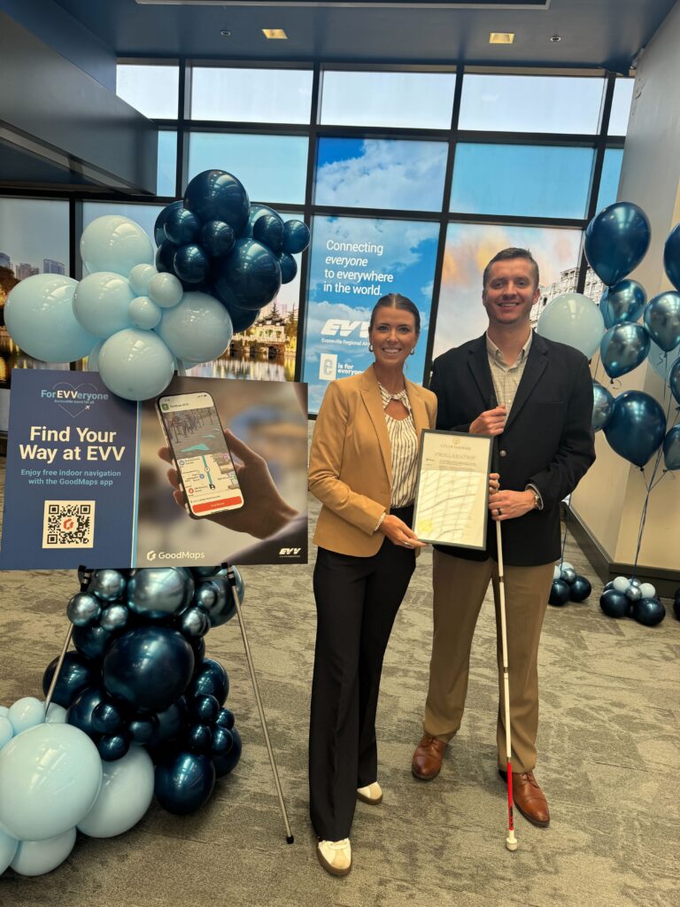 Evelyn and Kevin at Evansville Regional Airport stand in front of a GoodMaps ‘Find Your Way at EVV’ display with blue balloons, holding a framed document and a white mobility cane.